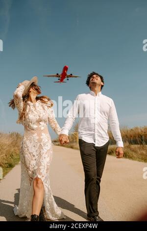 Glücklicher junger Mann in stilvollem Outfit, der die Hand der Frau im weißen Hochzeitskleid hält, während er mit fliegendem Flugzeug auf der Landstraße gegen den blauen Himmel schlendert Stockfoto