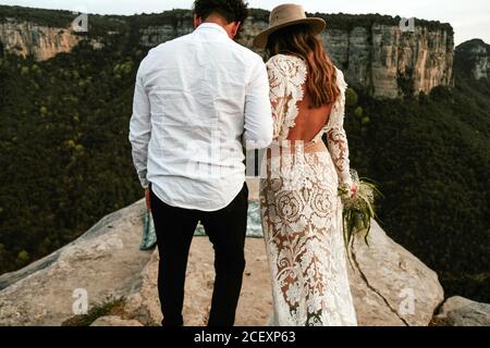 Rückansicht von unkenntlich newlywed Paar in eleganten trendigen Hochzeit Kleidung mit Blumenstrauß steht am Rand der felsigen Klippe während Besuch von Morro de Labella in Spanien Stockfoto