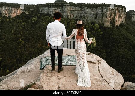 Ganzkörper-Rückansicht von unkenntlich Neuvermählten Paar in elegant Trendige Hochzeitskleidung mit Blumenstrauß am Rand von felsig stehen Klippe während eines Besuchs in Morro de Labella in Spanien Stockfoto