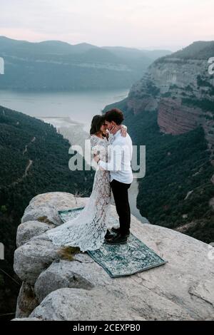 Seitenansicht eines jungen Mannes und einer Frau in eleganter Kleidung, die auf einer felsigen Klippe von Morro de Labella stehen und sich umarmen und sich sanft küssen Stockfoto