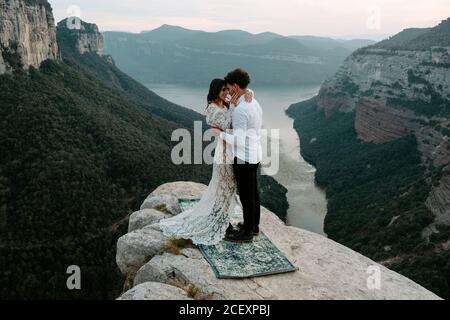 Seitenansicht eines jungen Mannes und einer Frau in eleganter Kleidung, die auf einer felsigen Klippe von Morro de Labella stehen und sich umarmen und sich sanft küssen Stockfoto