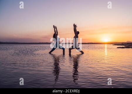 Volle Seitenansicht von unkenntlichen Männern und Frauen in Sportkleidung, die acroyoga praktizieren und Crescent Lunge Asana machen, während sie im Wasser des Sees gegen den Sonnenuntergang stehen Stockfoto