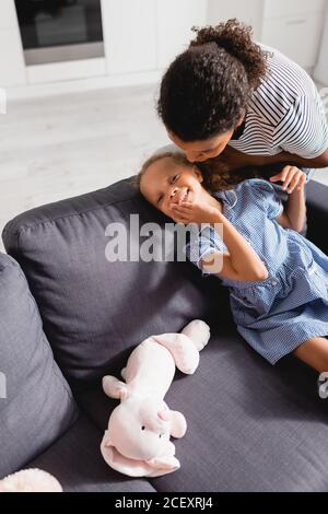 afroamerikanische Frau flüstert im Ohr der aufgeregt Tochter berühren Gesicht, während auf der Couch in der Nähe Spielzeug Hase sitzen Stockfoto