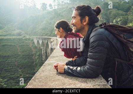 Seitenansicht eines fröhlichen ethnischen Mannes und einer Frau in aktiver Atmosphäre mit Rucksäcken, die die Aussicht auf die alte Nine Arch Bridge aus Stein zwischen grünen Hügeln in Sri Lanka betrachten Stockfoto