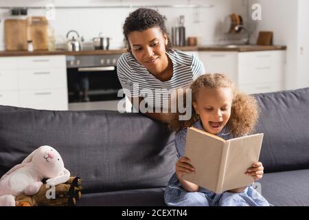 Junge afroamerikanische Babysitter hinter aufgeregt Kind reding Buch auf Couch in der Küche Stockfoto