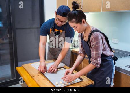 Junge asiatische Mann und Frau in Schürzen arbeiten zu Hause Bäckerei und setzen Cookies auf Backform mit Pergament Stockfoto