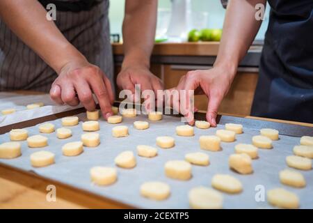 Zugeschnittene, nicht erkennbare Hände von Mann und Frau in Schürzen, die in der Bäckerei zu Hause arbeiten und Cookies auf Backform mit Pergament legen Stockfoto