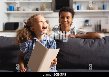 Selektive Fokus der afroamerikanischen Babysitter hinter aufgeregt Mädchen halten Öffnen Sie das Buch, während Sie auf dem Sofa sitzen Stockfoto