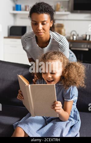 Junge afroamerikanische Babysitter im gestreiften T-Shirt hinter aufgeregt Mädchen Buch auf Sofa lesen Stockfoto