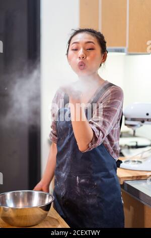 Charmante junge asiatische Frau in Schürze bläst Mehl Pulver stehen an Holztisch in der Küche Stockfoto