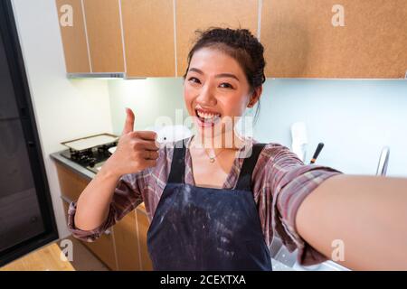 Glückliche asiatische Frau in unordentlicher Schürze zeigt Daumen vor der Kamera, während sie Selfie in der heimischen Küche macht Stockfoto