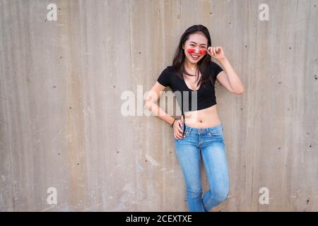 Entzückte junge asiatische Frau in stilvollem Outfit und Sonnenbrille lehnte Auf Steinmauer auf der Straße und Blick auf die Kamera Stockfoto