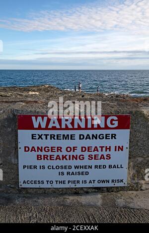 Ein Schild, das vor den Gefahren von Wellen und Gezeiten an einem Strand in Cornwall, England, warnt, mit Menschen auf den Felsen am Ufer. Stockfoto