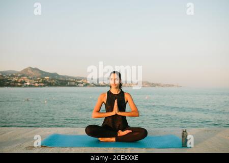 Entspannte Frau sitzt auf Yogamatte in Padmasana mit Mudra Hände und Übung Achtsamkeit Blick weg auf den Hintergrund der Seeslandschaft Bei Sonnenuntergang Stockfoto