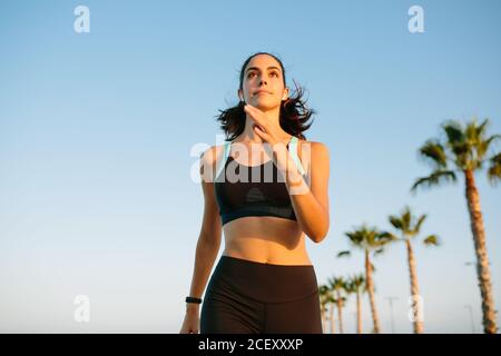 Von unten bestimmt junge schlanke Weibchen im schwarzen Sport Top und Leggings laufen auf der Straße mit Palmen gegen Blauer Himmel Stockfoto