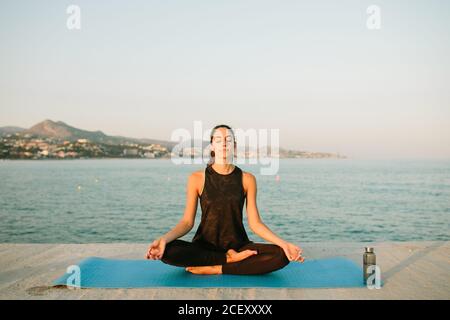 Entspannte Frau sitzt auf Yogamatte in Padmasana mit Mudra Hände und Übung Achtsamkeit mit geschlossenen Augen auf Hintergrund seascape bei Sonnenuntergang Stockfoto