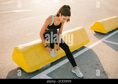 Ganzkörper der jungen schlanken Läuferin in schwarzer Sportbekleidung Sitzen auf Betongrenze auf gepflasterten Straße und binden Schnürsenkel Auf Sneakers während der Vorbereitung auf das Joggen Stockfoto