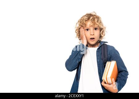 Schockiert blonde Schuljunge hält Bücher und berühren Gesicht isoliert auf Weiß Stockfoto