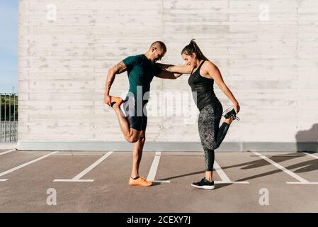 Seitenansicht eines starken Sportlers und einer Sportlerin, die sich aufeinander lehnen und die Beine strecken, bevor sie an sonnigen Tagen in der Stadt trainieren Stockfoto