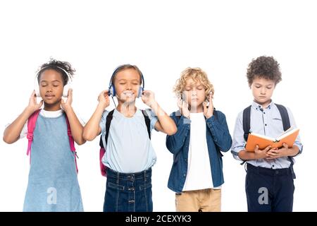 Multikulturelle Schüler hören Musik in kabellosen Kopfhörern in der Nähe von lockigen Jungen Lesebuch isoliert auf weiß Stockfoto