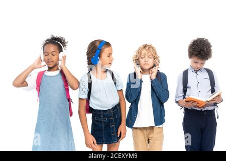 Schulmädchen Musik in kabellosen Kopfhörern hören und Blick auf lockig Junge Buch bei multikulturellen Schulkindern isoliert auf weiß lesen Stockfoto