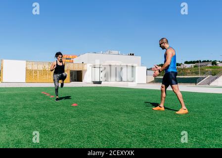 Ganzkörper junge Athletin springen auf Agility Leiter während Fitnesstraining mit Personal Trainer auf Sportplatz Stockfoto