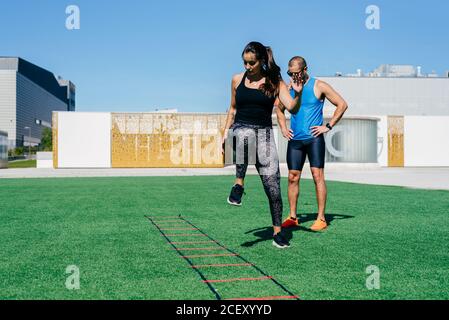 Ganzkörper junge Athletin springen auf Agility Leiter während Fitnesstraining mit Personal Trainer auf Sportplatz Stockfoto