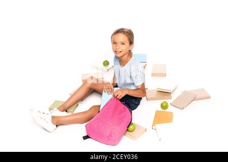 Schulmädchen sitzt in der Nähe von Äpfeln und Putting Buch im Rucksack isoliert Auf Weiß Stockfoto