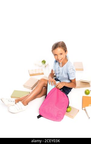 Schulkind sitzt in der Nähe von Äpfeln und Putting Buch in Rucksack isoliert Auf Weiß Stockfoto