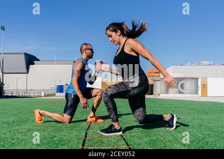 Ganzkörper-Seitenansicht von fokussierten, sportlichen Männern und Frauen, die beim Training mit einer Agility-Leiter auf dem grünen Rasen auf dem Sportplatz Ausfallschritte machen Stockfoto
