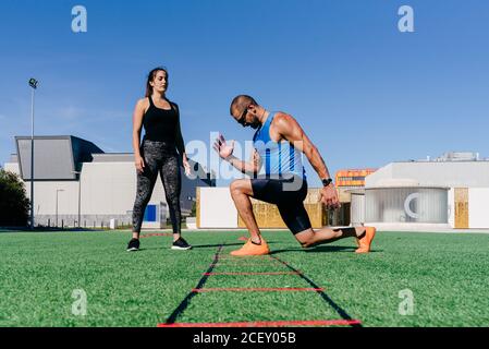 Ganzkörper-Seitenansicht des fokussierten sportlichen Mannes beim Ausfallschritt Beim Training mit Agility Leiter auf grünen Rasen auf Sportplatz Stockfoto