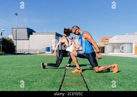 Ganzkörper-Seitenansicht von fokussierten, sportlichen Männern und Frauen, die beim Training mit einer Agility-Leiter auf dem grünen Rasen auf dem Sportplatz Ausfallschritte machen Stockfoto