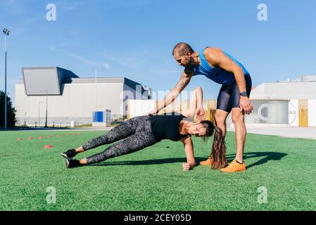 Ganzkörper eines männlichen persönlichen Trainers, der jungen Frau bei der Durchführung von Seitenplankenübungen beim Fitnesstraining auf der Stadionwiese hilft Stockfoto