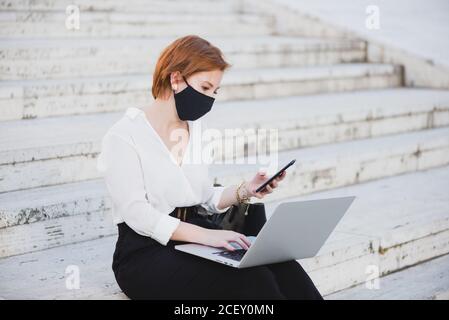 Konzentrierte Geschäftsfrau in eleganter Kleidung und medizinischer Maske sitzt auf der Straße auf Treppen und arbeitet an Remote-Projekt, während mit Laptop und Smartphone Stockfoto
