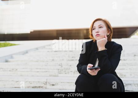 Durchdachte Managerin in elegantem Anzug, die Treppen in der Stadt sitzt Und auf dem Handy, während Sie wegschauen Stockfoto