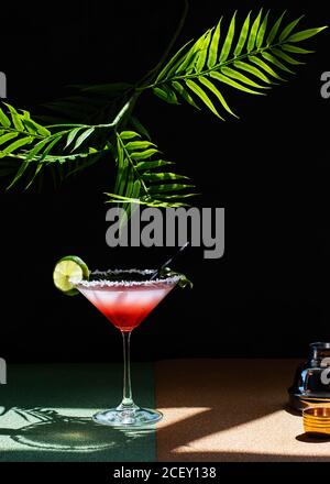 Tropischer Margarita Cocktail mit Grenadine auf glitzerndem Tisch platziert mit Pflanzenblätter auf schwarzem Hintergrund Stockfoto