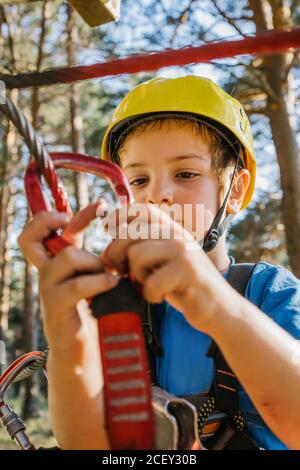 Entzückendes Kind in Schutzhelm und Sicherheitsgurt Putting Karabine Am Seil, während Sie im Sommer ein Wochenende im Abenteuerpark verbringen Stockfoto