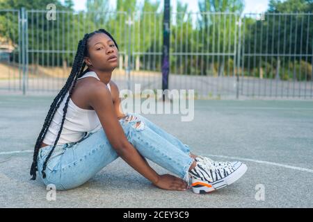 Volle Länge der jungen afroamerikanischen Frau mit Zöpfen tragen Trendige zerrissene Jeans und weißes Tank-Top mit Sneakers sitzen Gegen Basketballkorb auf dem Spielplatz und Blick auf die Kamera Stockfoto