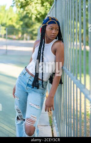 Junge afroamerikanische Hündin mit Afro-Zöpfen, die stilvoll gekleidet sind Zerrissene Jeans und weißes Oberteil, das auf Metallzaun lehnt Stadtpark und Blick weg Stockfoto