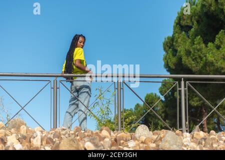 Von unten Seitenansicht einer selbstbewussten jungen afroamerikanischen Frau Hipster in buntem T-Shirt und Jeans in der Nähe von Banister stehen Im Sommer Park und Blick auf die Kamera Stockfoto