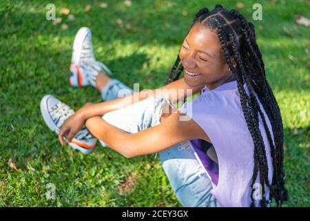 Von oben gesehen, die glückliche junge afroamerikanische Frau mit Zöpfen in legerer Kleidung auf grünem Gras sitzend und lachend, während sie den Sommertag im Park genießt Stockfoto