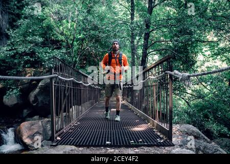 Sorgloser männlicher Tourist mit Rucksack, der entlang einer Hängebrücke aus Metall läuft Und bewundern erstaunliche Natur der Wälder während des Urlaubs Stockfoto