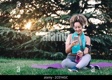 Ganzkörper fröhlich junge lockig behaarte Weibchen in Sportswear mit Kopfhörer sitzen auf Gras und browsen Smartphone während der Ruhe mit Flasche Wasser nach dem Training im Sommerpark Stockfoto