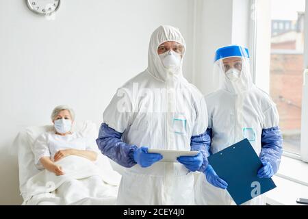 Horizontales Mittelportrait von zwei modernen Ärzten, die zusammen stehen Krankenstation mit Patient hinter ihnen und Blick auf die Kamera Stockfoto