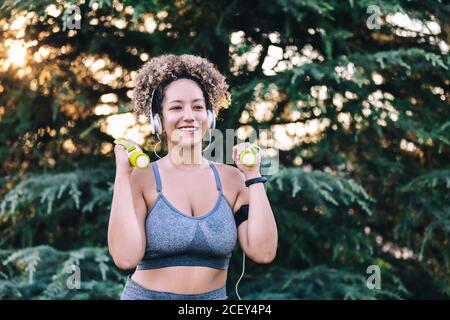 Junge ethnisch lockig haarige Frau in Sport-Top und Leggings Mit Hanteln trainieren und mit Kopfhörern Musik hören Beim Arbeiten im grünen Park Stockfoto