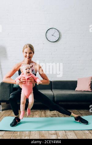Sportlerin dabei Ausfallschritte, während Baby Mädchen auf Fitness-Matte halten Zu Hause Stockfoto
