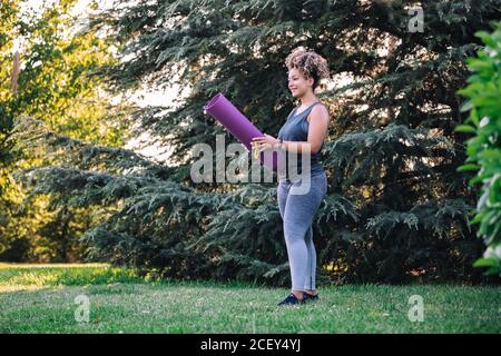 Ganzkörper Seitenansicht der fröhlichen jungen ethnischen weiblich in activewear hält die Rollmatte und bereitet sich auf das Training auf Grün vor Rasen im Park Stockfoto