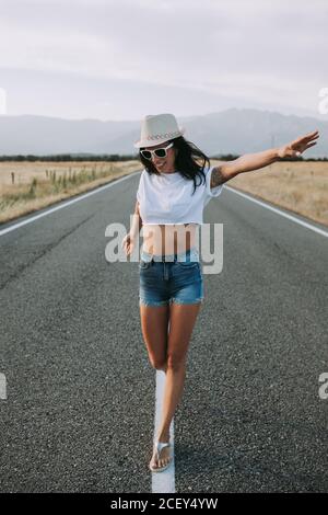 Ruhige weibliche Reisende im Sommer tragen Wandern mit ausgestreckten Armen Entlang der leeren Asphaltstraße auf dem Hintergrund der bergigen Landschaft und Genießen Sie die Freiheit Stockfoto