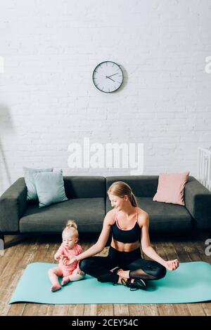 Mutter in Sportkleidung sitzt in Yoga-Pose in der Nähe von Kleinkind Tochter Auf der Fitnessmatte Stockfoto