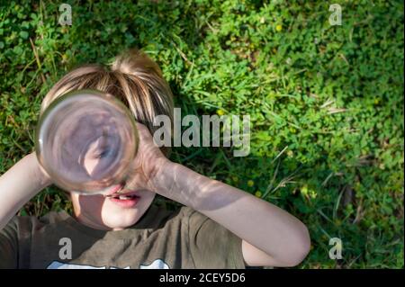 Von oben entzückendes Kind, das im Sommer auf Gras liegt Und die Kamera durch die Flasche betrachten, so als ob sie mit Spyglass spielt Stockfoto